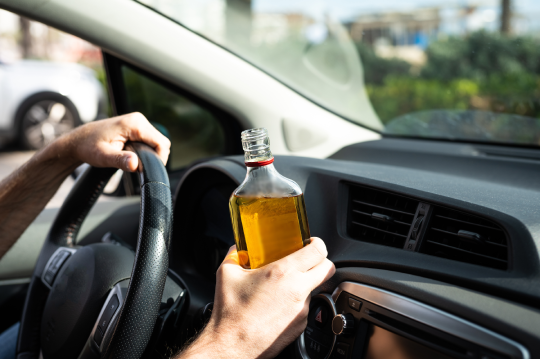 Man holding a bottle of alcohol while driving a car, highlighting the danger and irresponsibility of drinking and driving.