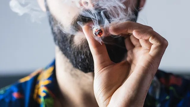 A close-up view of a bearded man wearing a colorful shirt, holding a lit marijuana joint to his lips and exhaling smoke.