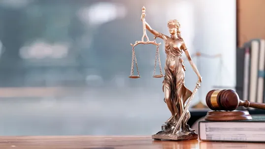 A bronze Lady Justice statue holding the scales of justice and a sword, standing on a wooden surface next to a law book and a wooden gavel, with a softly blurred background.