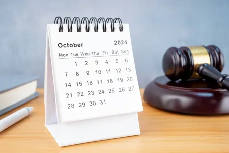 A spiral-bound desktop calendar open to October 2024 sitting on a wooden desk next to a wooden legal gavel and sound block.