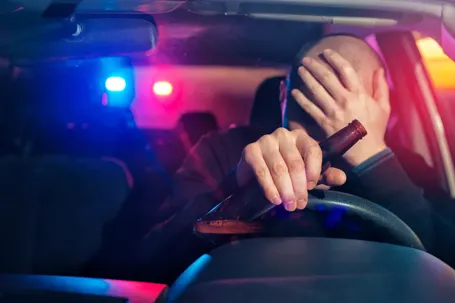 An interior shot from a car showing a driver holding an open beer bottle and covering their face in distress, with the flashing red and blue lights of a police car visible through the rear window.