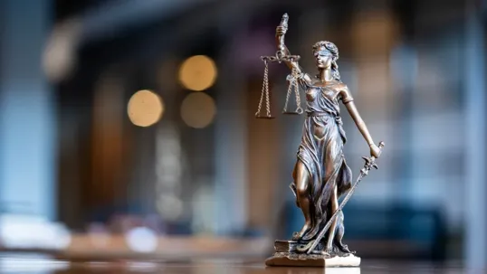 A bronze Lady Justice statue holding the scales of justice and a sword, standing on a wooden surface with a softly blurred, warm-toned office interior in the background.