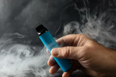 A close-up shot of a person's hand holding a bright blue disposable vape pen surrounded by thick plumes of white vapor against a dark, textured background.